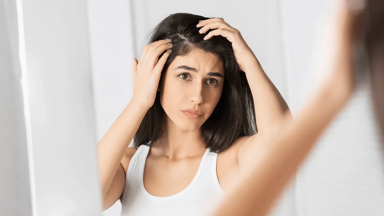 A standing woman looking in a mirror while checking the roots of her long, dark hair.