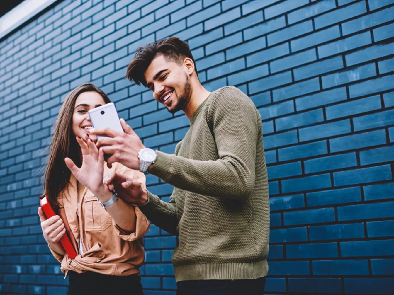 Two people looking at a cell phone while standing in front of a blue brick wall.